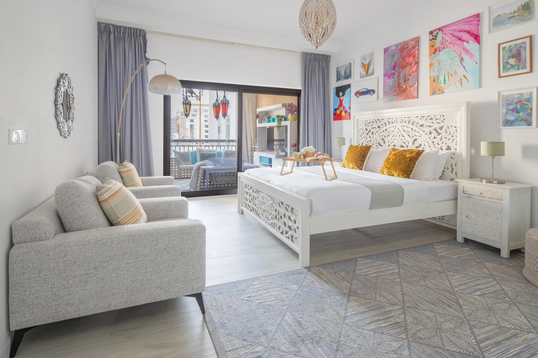 Un dormitorio luminoso con una cama blanca, asientos grises y coloridos cuadros.