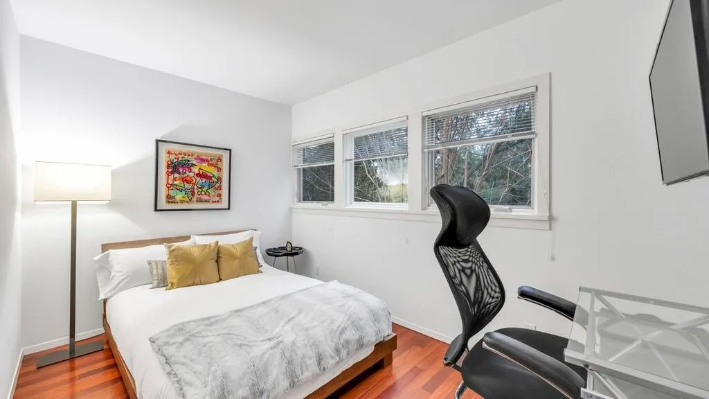 Un dormitorio con una cama, un cuadro enmarcado y una silla de escritorio junto a las ventanas.