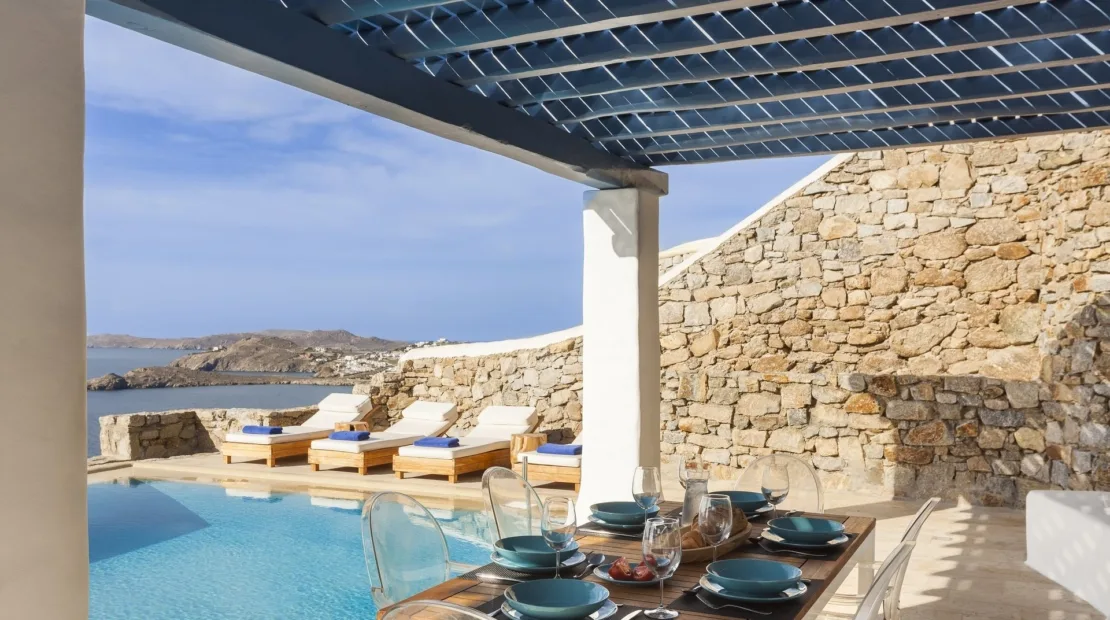 A covered outdoor dining area overlooks a pool, loungers, and stone walls.