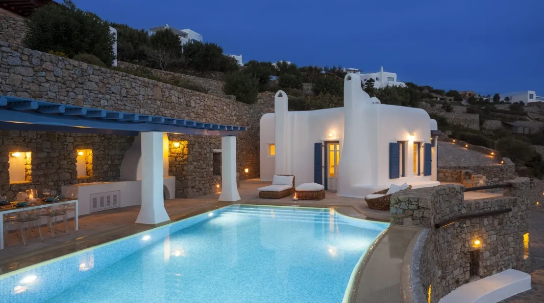 A swimming pool beside a white villa, with stone terraces and outdoor seating.