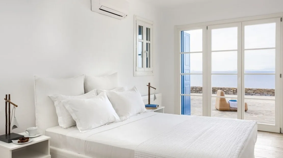 A bright bedroom with a white bed beside glass doors opening onto a terrace.