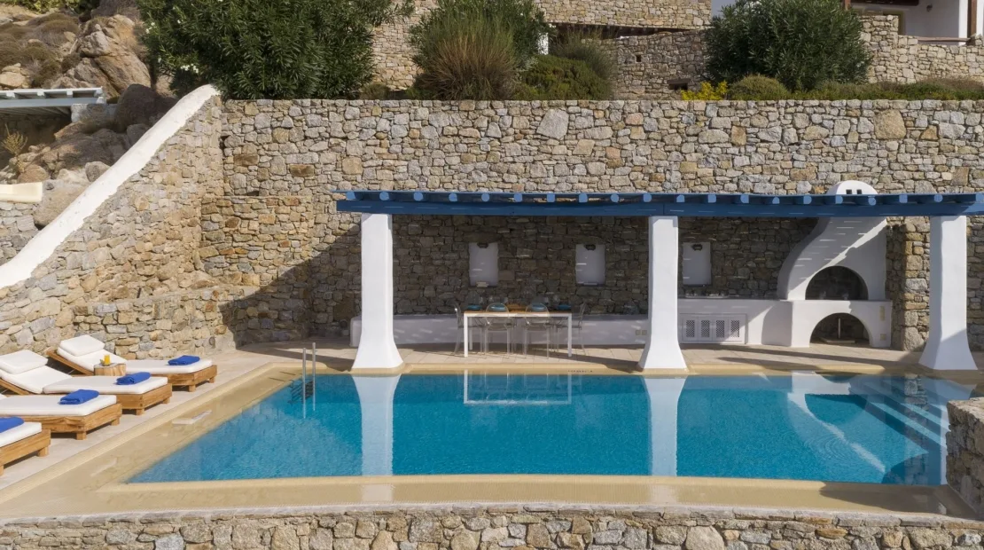 An outdoor pool sits beside a stone wall and shaded seating area.