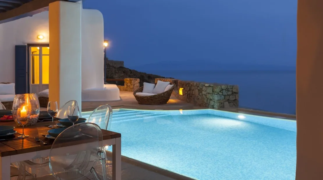 A lit swimming pool beside an outdoor dining area overlooks the sea.