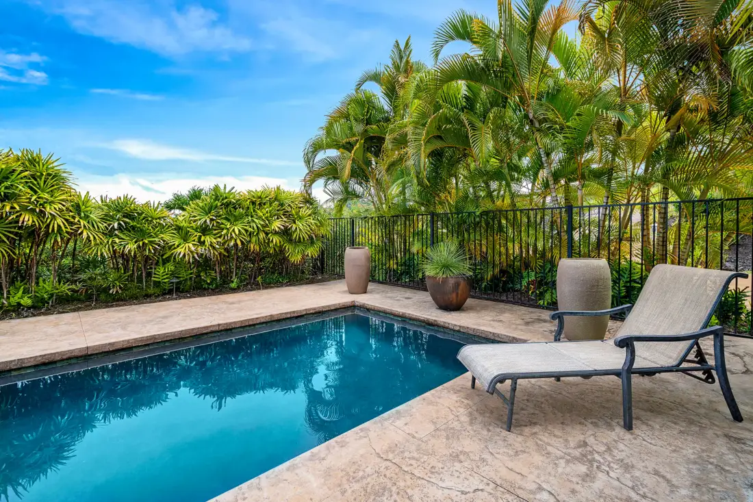 Una piscina al aire libre junto a una terraza pavimentada con asientos y plantas tropicales.