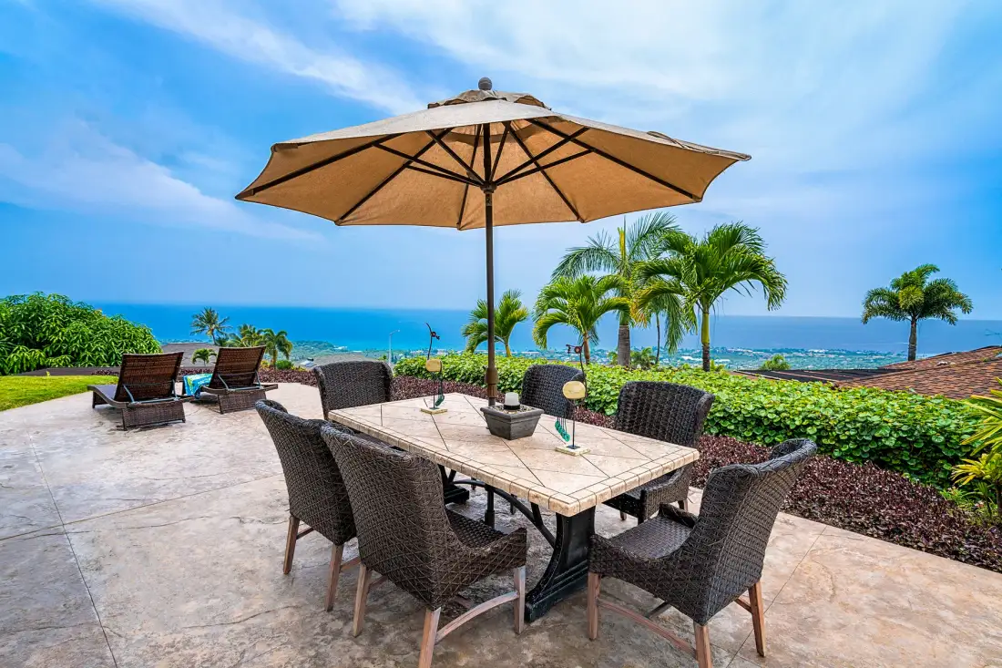 Una zona de comedor al aire libre con mesa y asientos bajo una gran sombrilla, frente al mar.