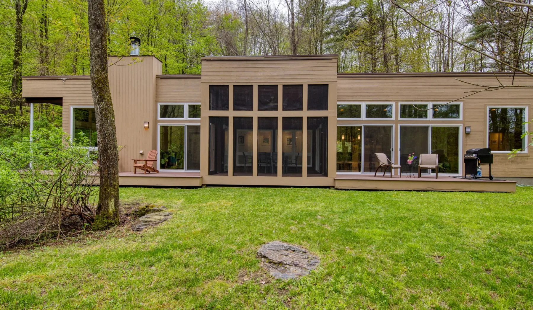 A modern house with broad windows faces a green lawn and surrounding trees.