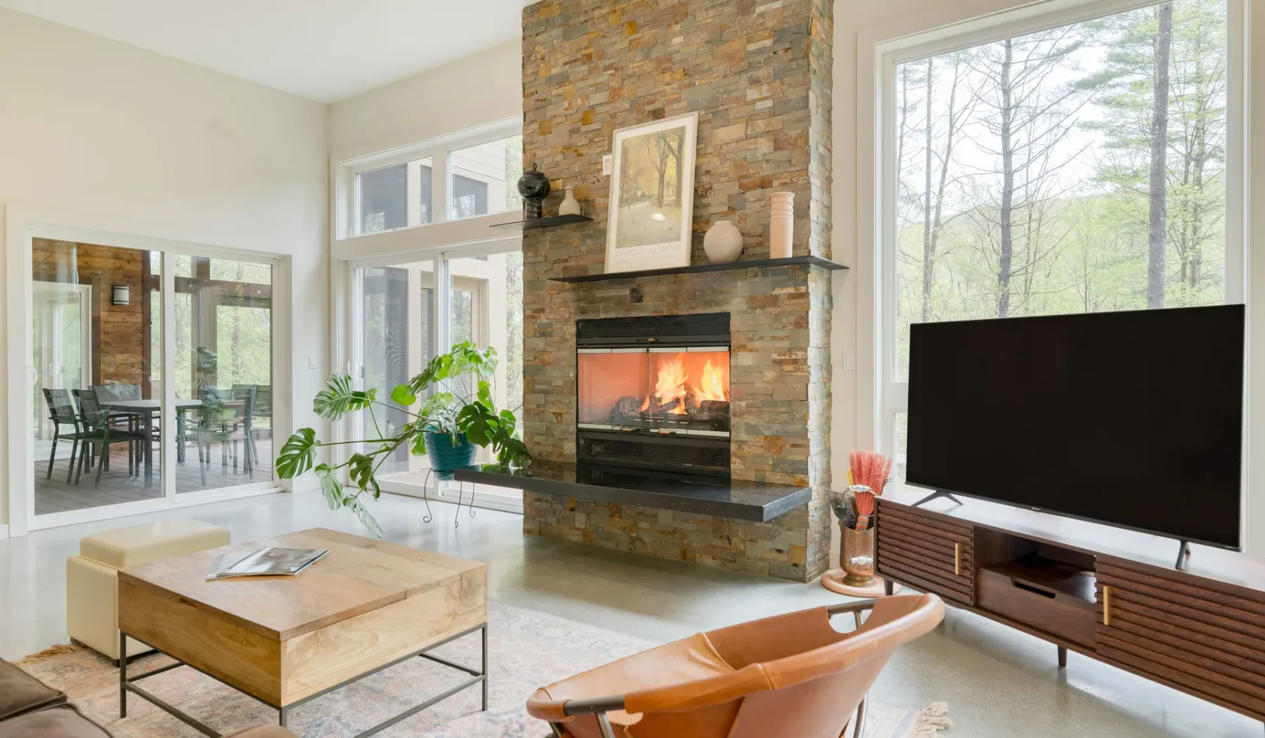 A bright living room features a stone fireplace, large windows, and a television.