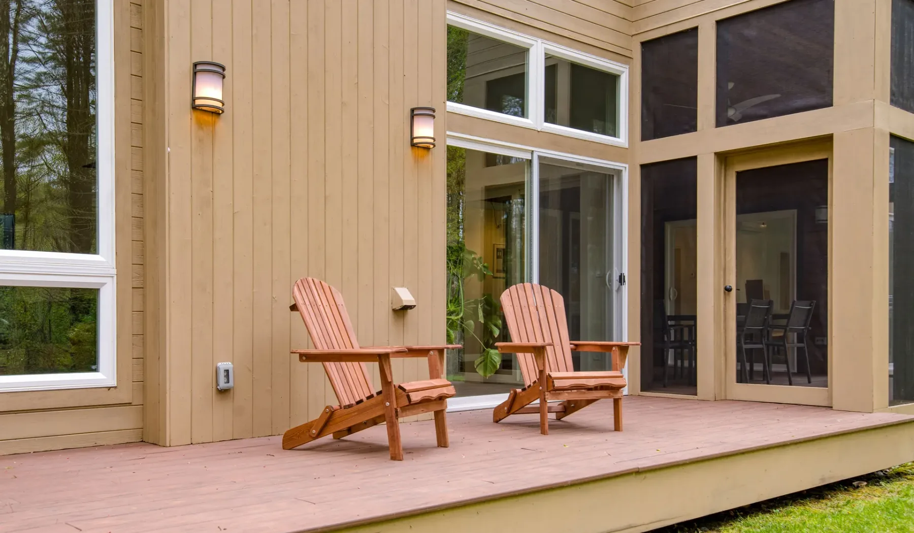 A wooden deck beside a modern home features two outdoor chairs and large windows.