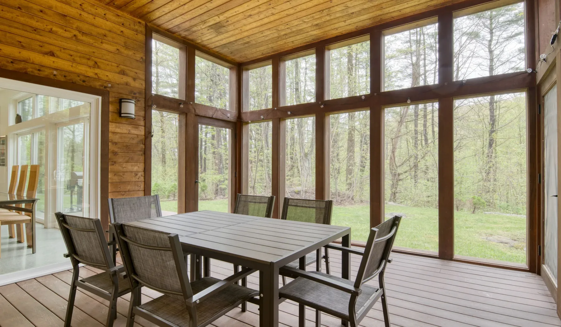 A dining table and chairs sit in a sunroom with large windows overlooking trees.