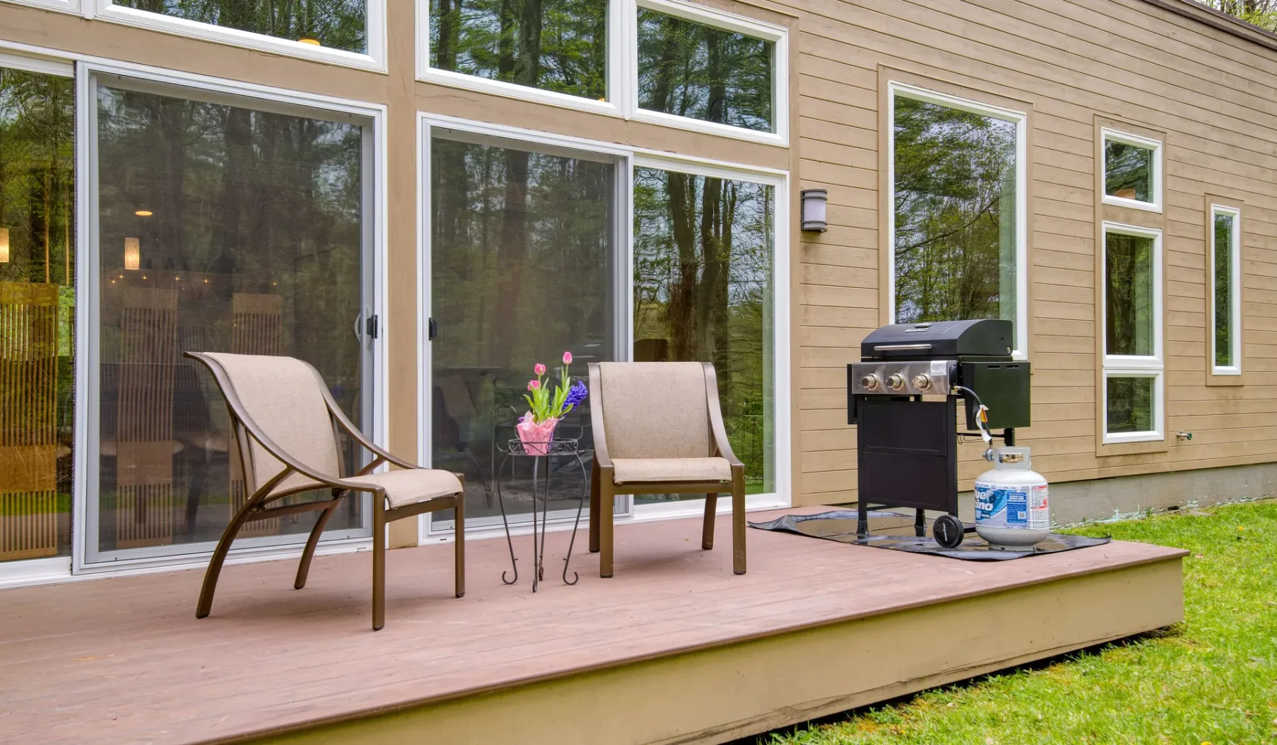 A wooden deck beside a house has two chairs, flowers, and an outdoor grill.