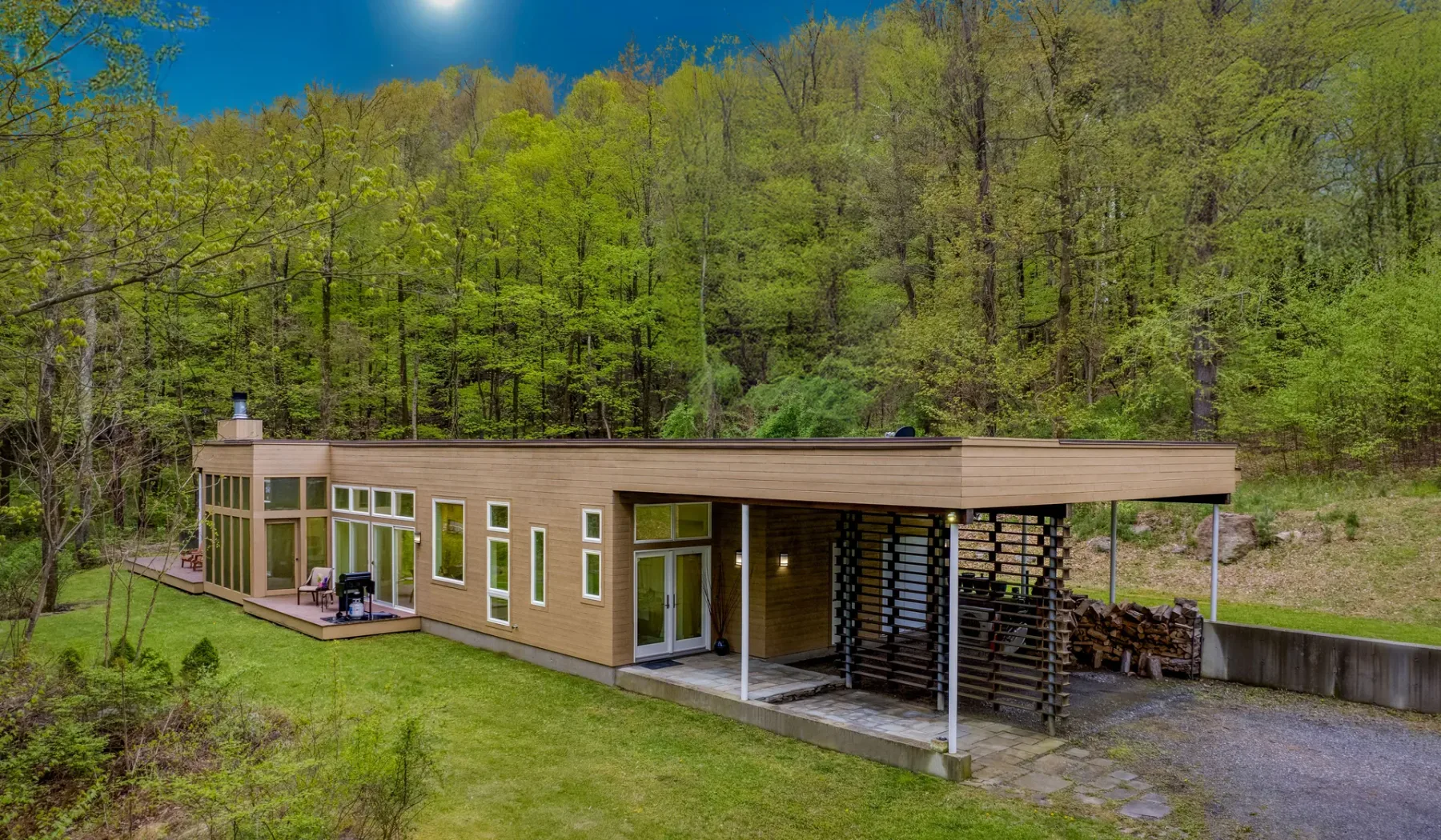 A modern wooden house sits beside a wooded hillside and gravel driveway.