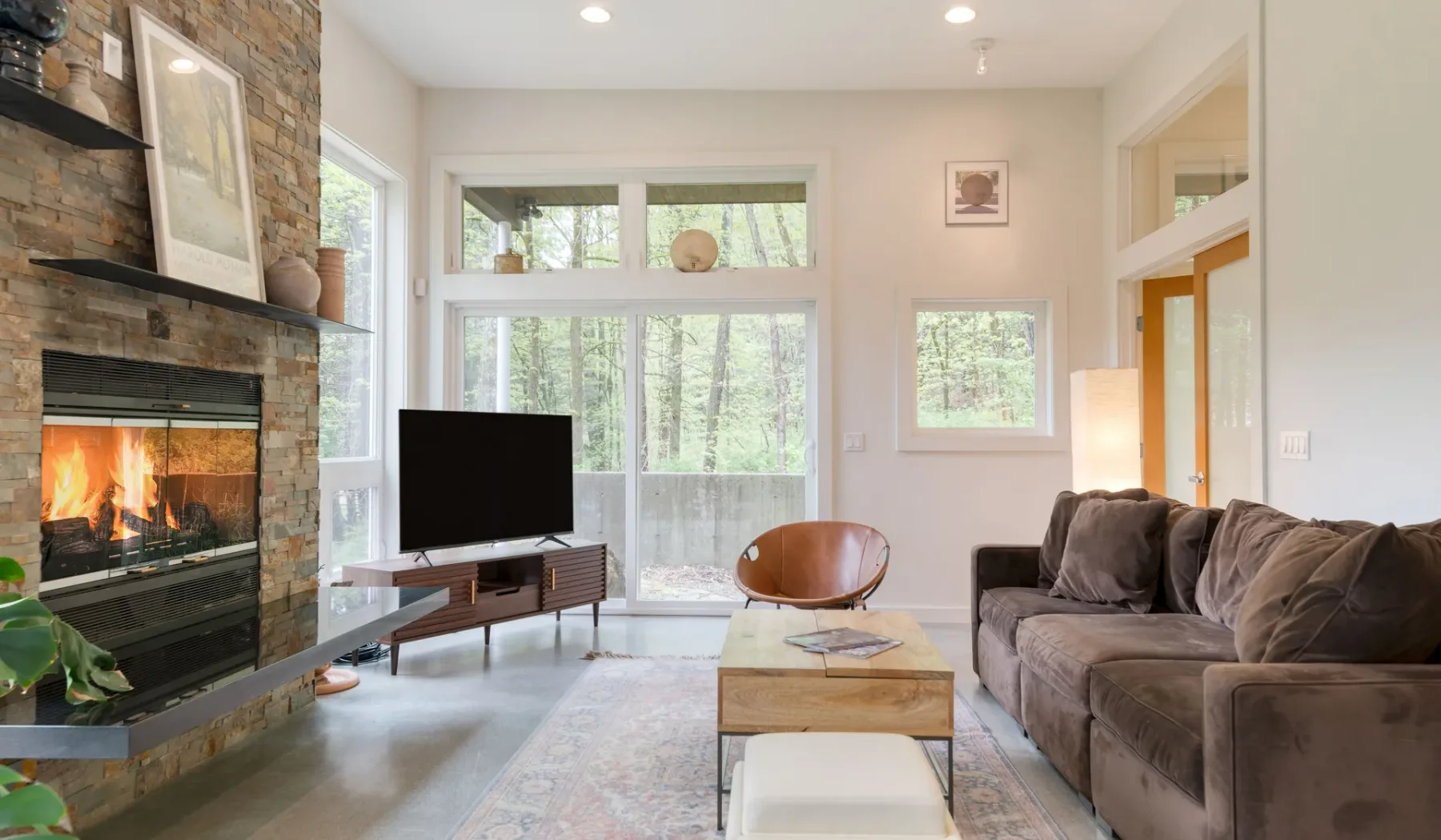 A living room with a stone fireplace, seating, and large windows overlooking trees.