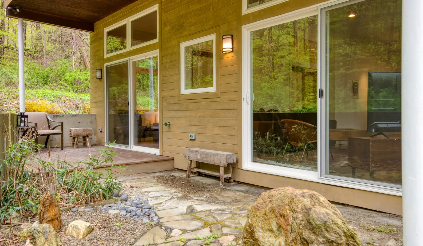A covered patio beside a wood-sided house, with large glass doors and stone paving.