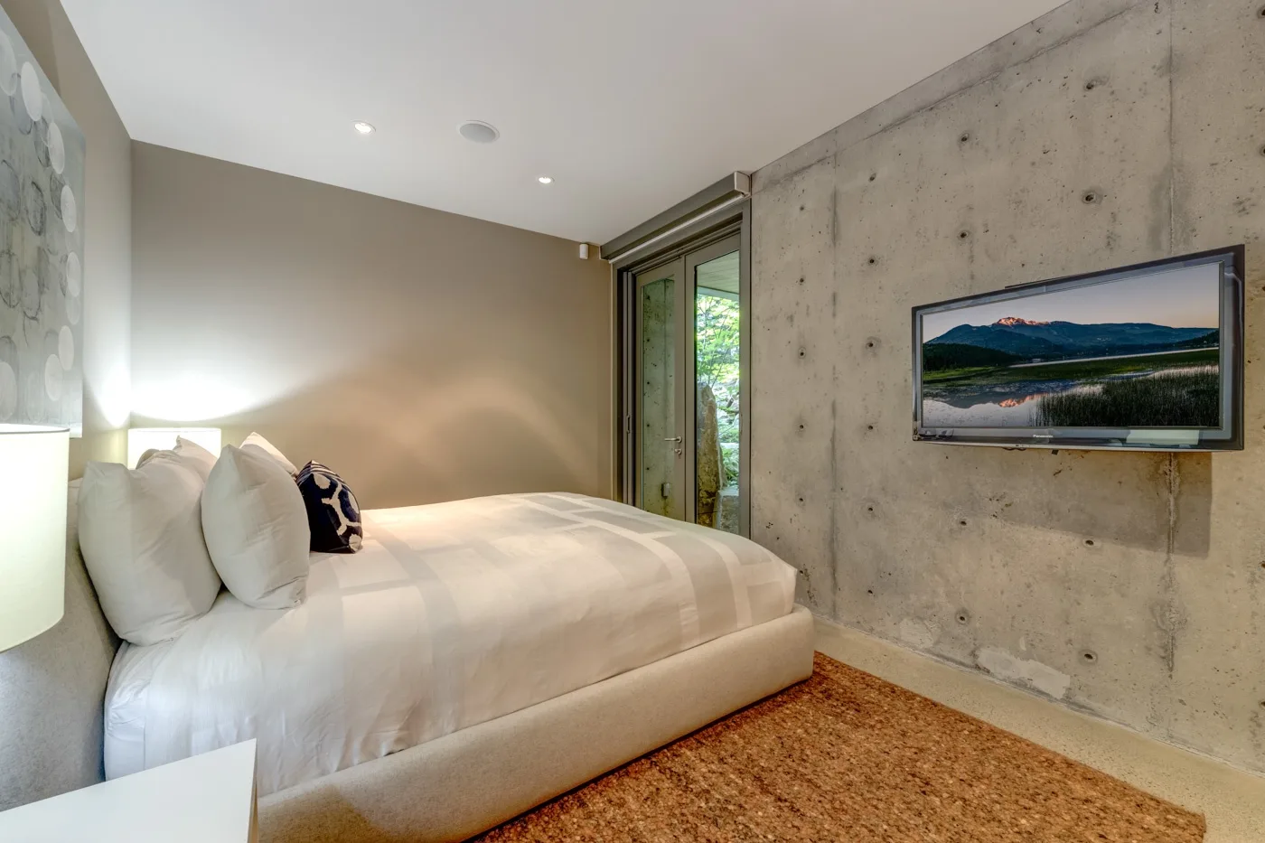 Un dormitorio con una cama junto a un televisor montado en la pared y ventanas altas.