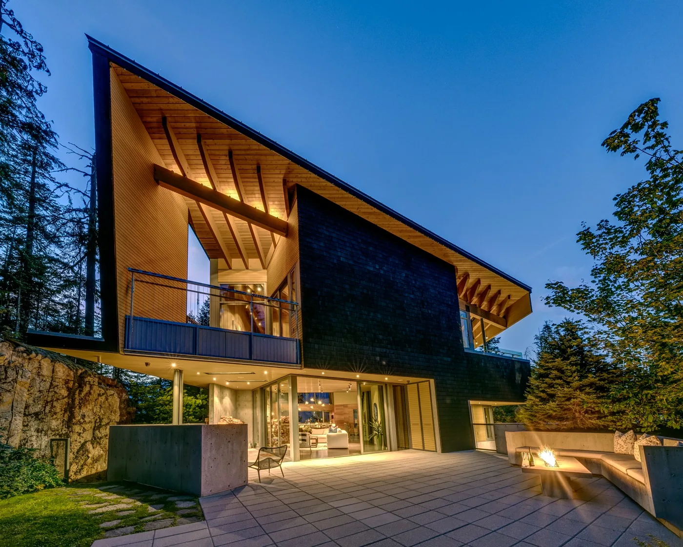 Una casa moderna al atardecer, con un amplio patio pavimentado y luces interiores cálidas.