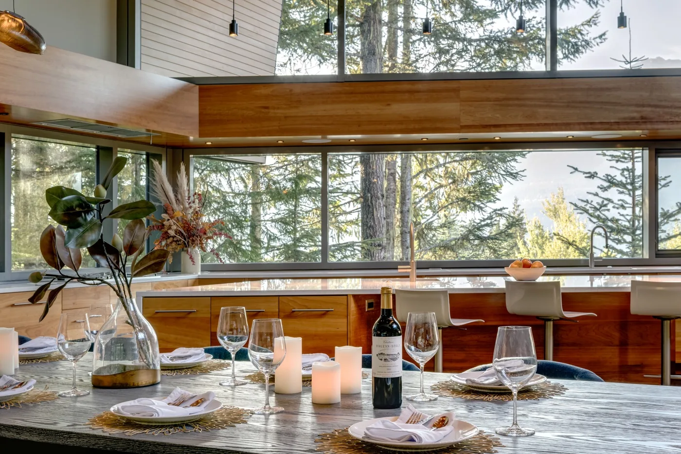 Una mesa está puesta en una habitación luminosa con grandes ventanas que dan a los árboles.