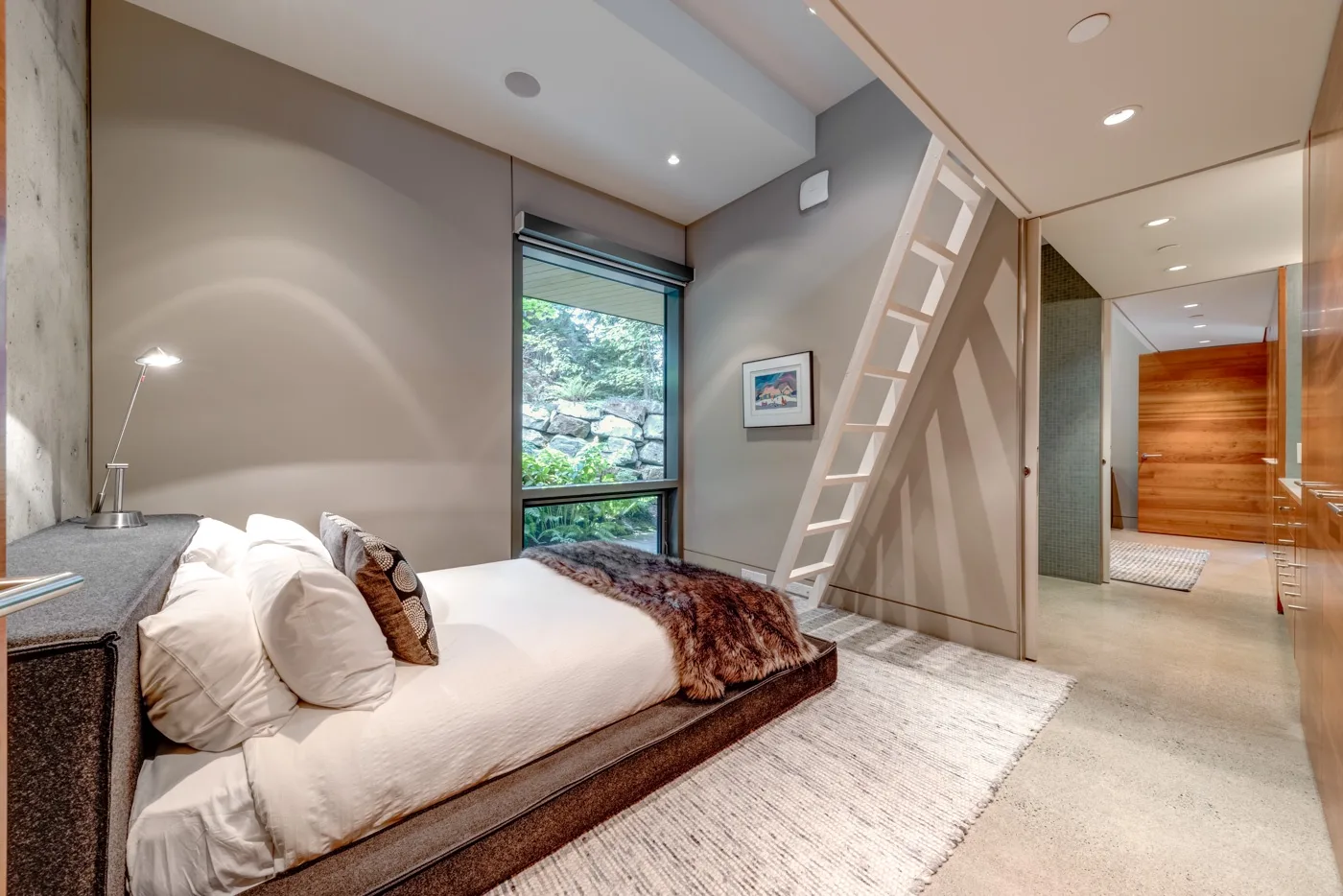 Un dormitorio con una cama baja junto a una ventana grande y una escalera hacia un altillo.