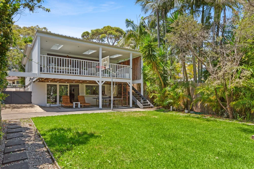 A raised house with a balcony overlooks a green lawn and tropical trees.