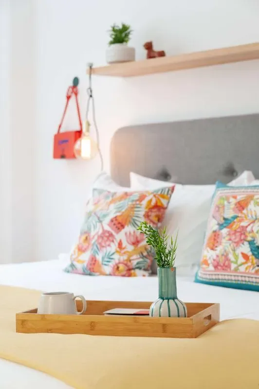 A neatly made bed has a gray upholstered headboard, colorful patterned pillows, and a wooden tray with a cup.