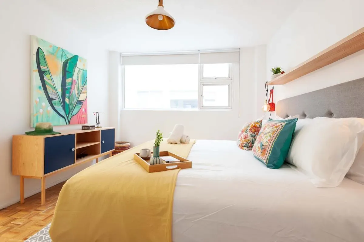 A bright bedroom features a white bed with a mustard throw, colorful pillows, and a wooden sideboard.