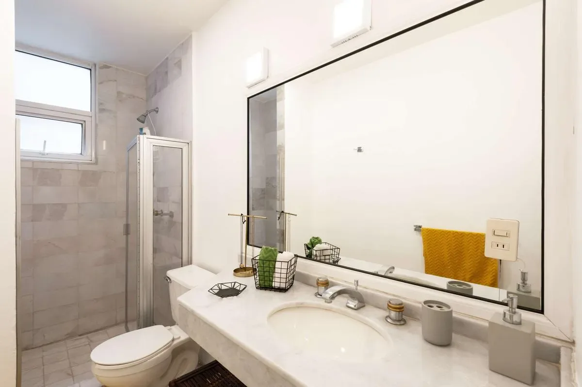 A bright bathroom features a long white vanity, large mirror, glass shower enclosure, and toilet.