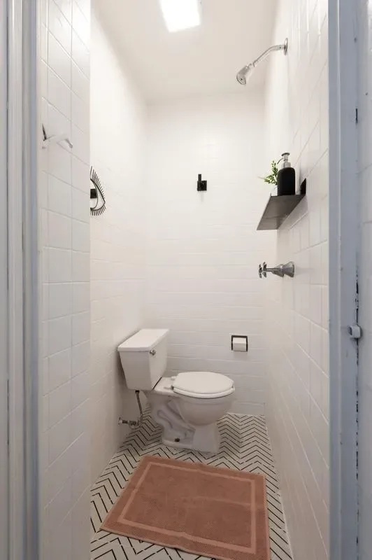 A narrow white-tiled bathroom has a toilet, wall-mounted shower fixtures, and a patterned floor with a brown bath mat.