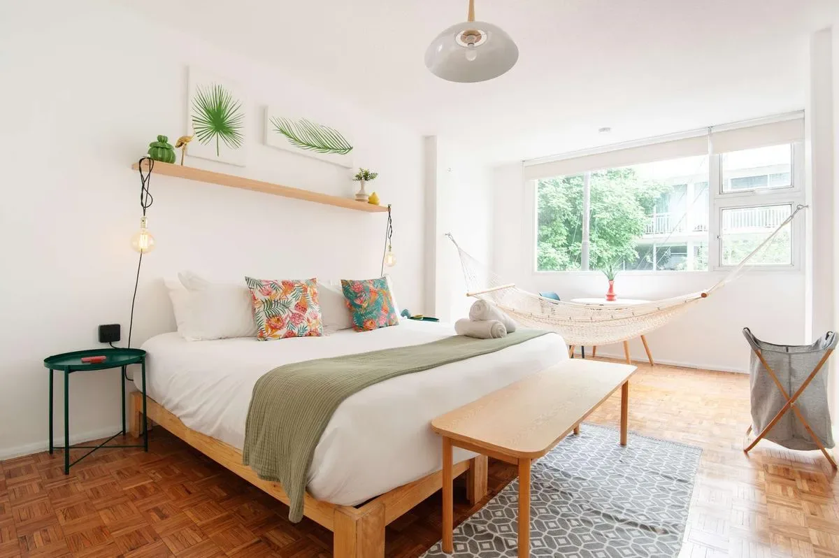 A bright bedroom features a low wooden bed, colorful cushions, a window hammock, and large windows.