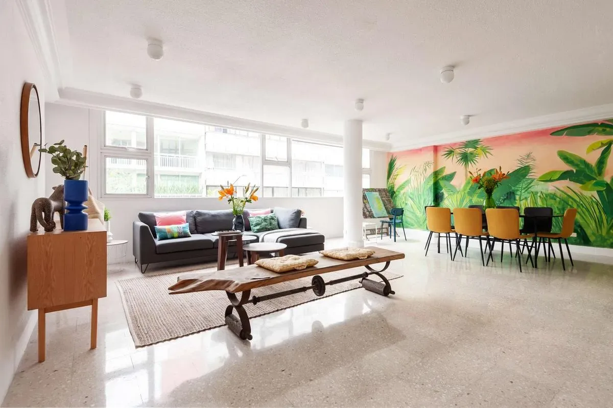A bright living room features a gray sofa, low wooden tables, and a colorful tropical mural beside a dining area.