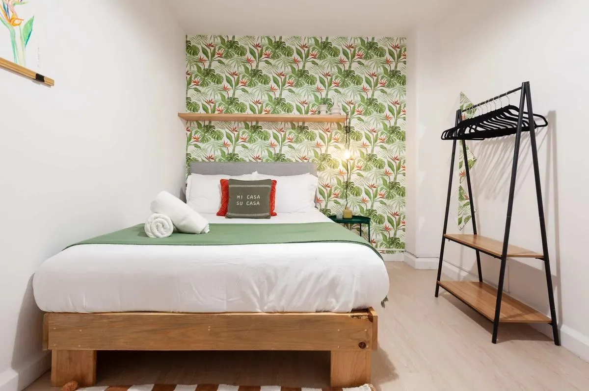 A neatly made bed with green bedding sits against a leafy patterned wall beside an open clothes rack.