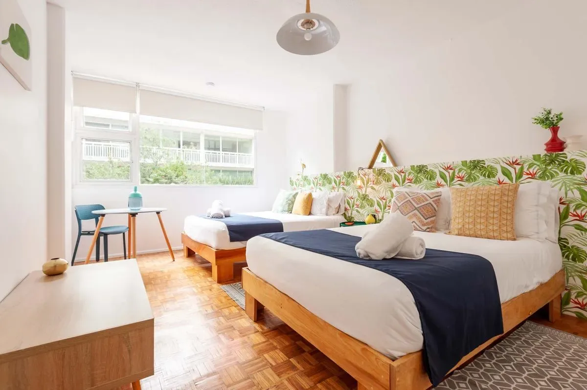A bright bedroom with two wooden beds, navy runners, patterned pillows, and a large window.