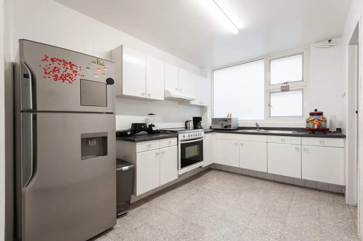 A bright kitchen has white cabinets, a stainless steel refrigerator, and a long countertop beneath two windows.