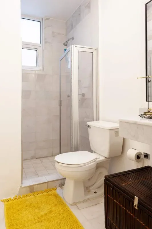 A compact tiled bathroom has a glass shower enclosure, toilet, wall-mounted sink, and yellow bath mat.