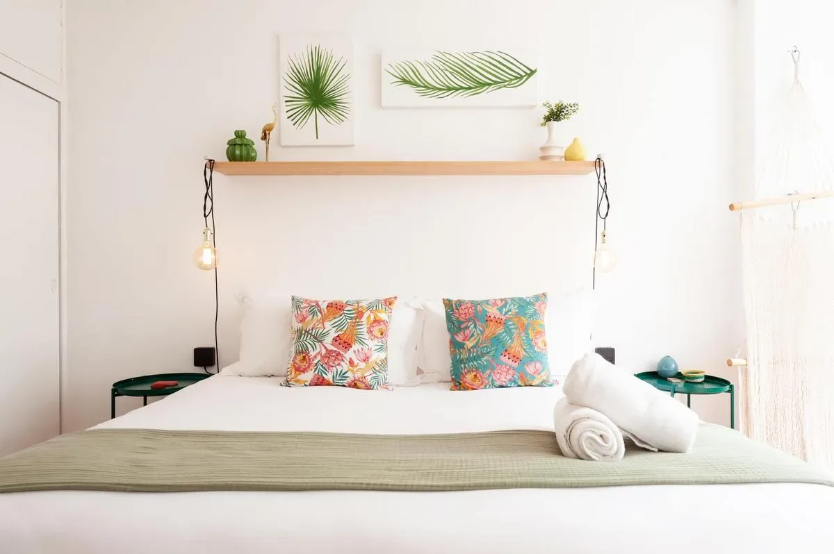 A neatly made bed has colorful floral pillows, a folded towel, and a wooden shelf with small plants above it.
