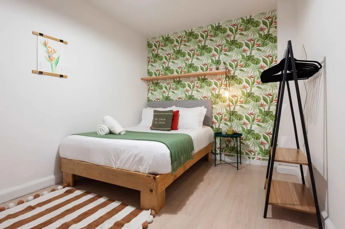 A bedroom with a wooden bed, green bedding, leafy wallpaper, and a freestanding clothes rack.