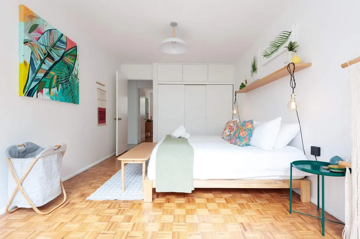 A bright bedroom with a white bed, colorful wall art, a wooden bench, and parquet flooring.