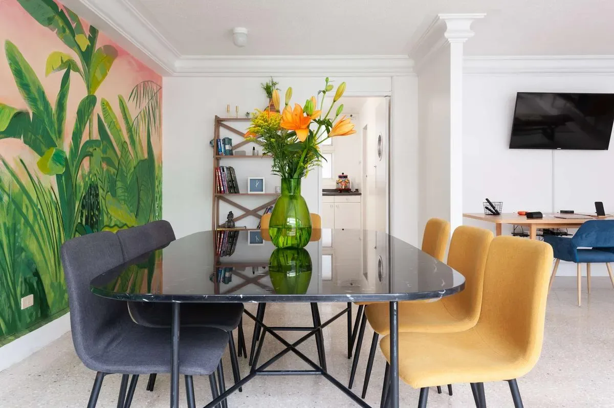 A dining table with a flower vase sits beside a colorful mural, with a television and desk in the background.