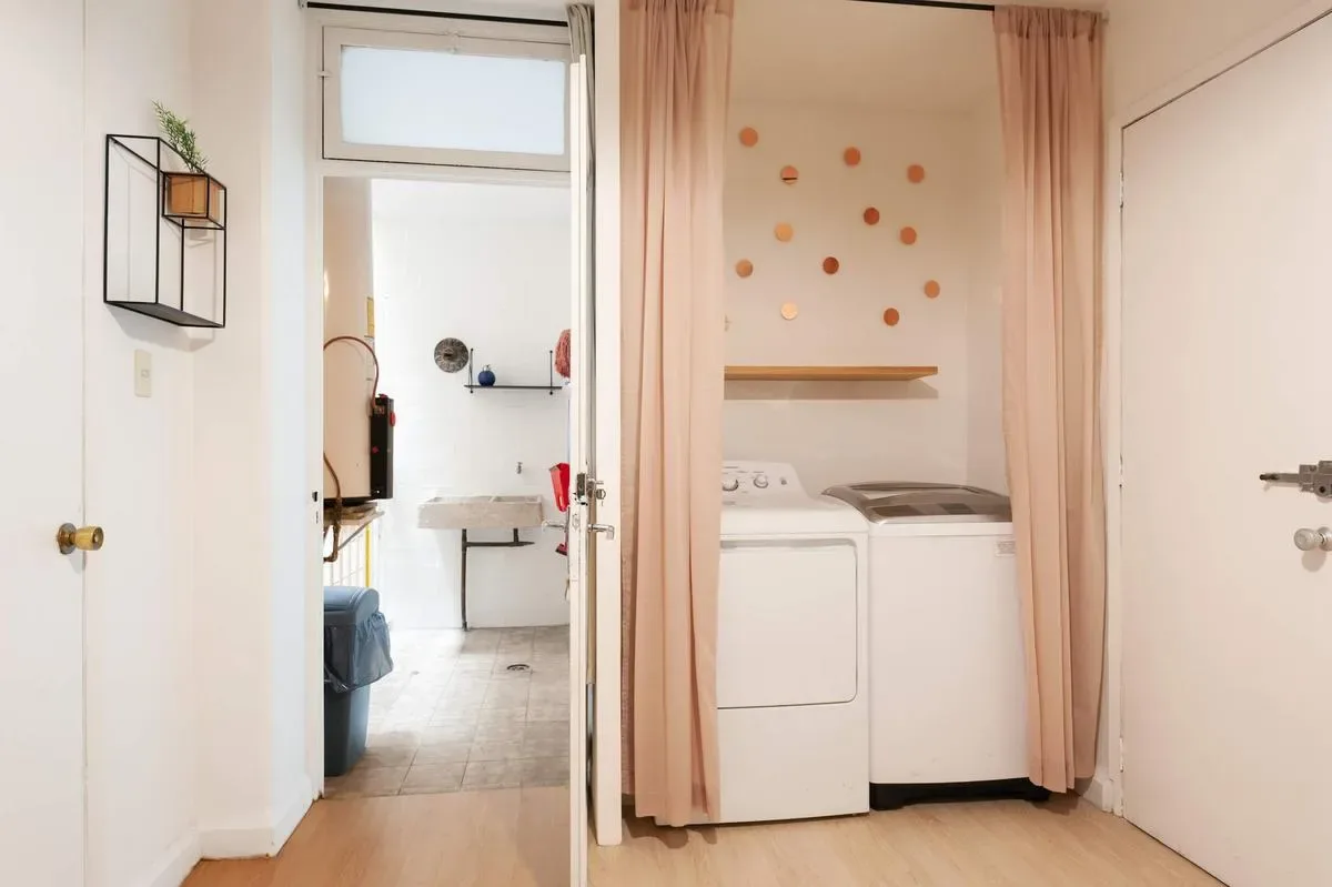 A compact laundry area has a washer and dryer behind pink curtains, with a bathroom visible through the doorway.