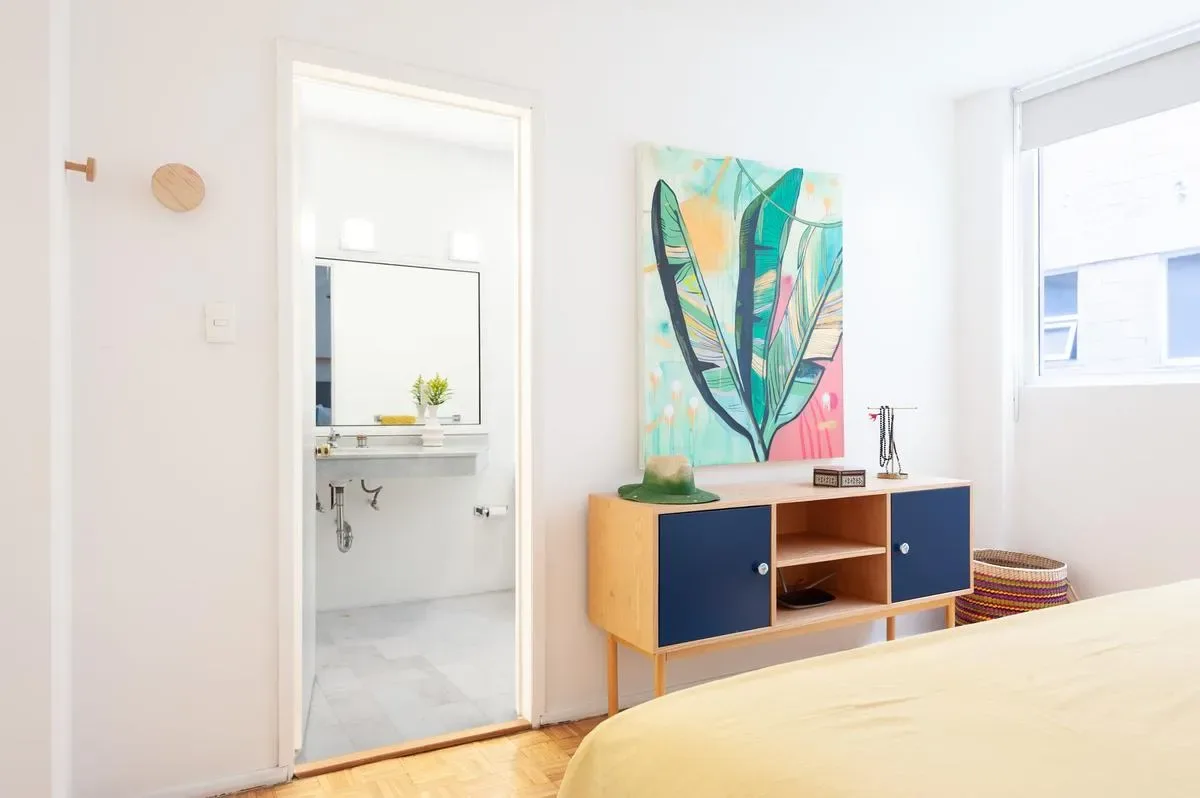 A bright bedroom with a low blue-and-wood cabinet, colorful abstract artwork, and a bed beside a large window.