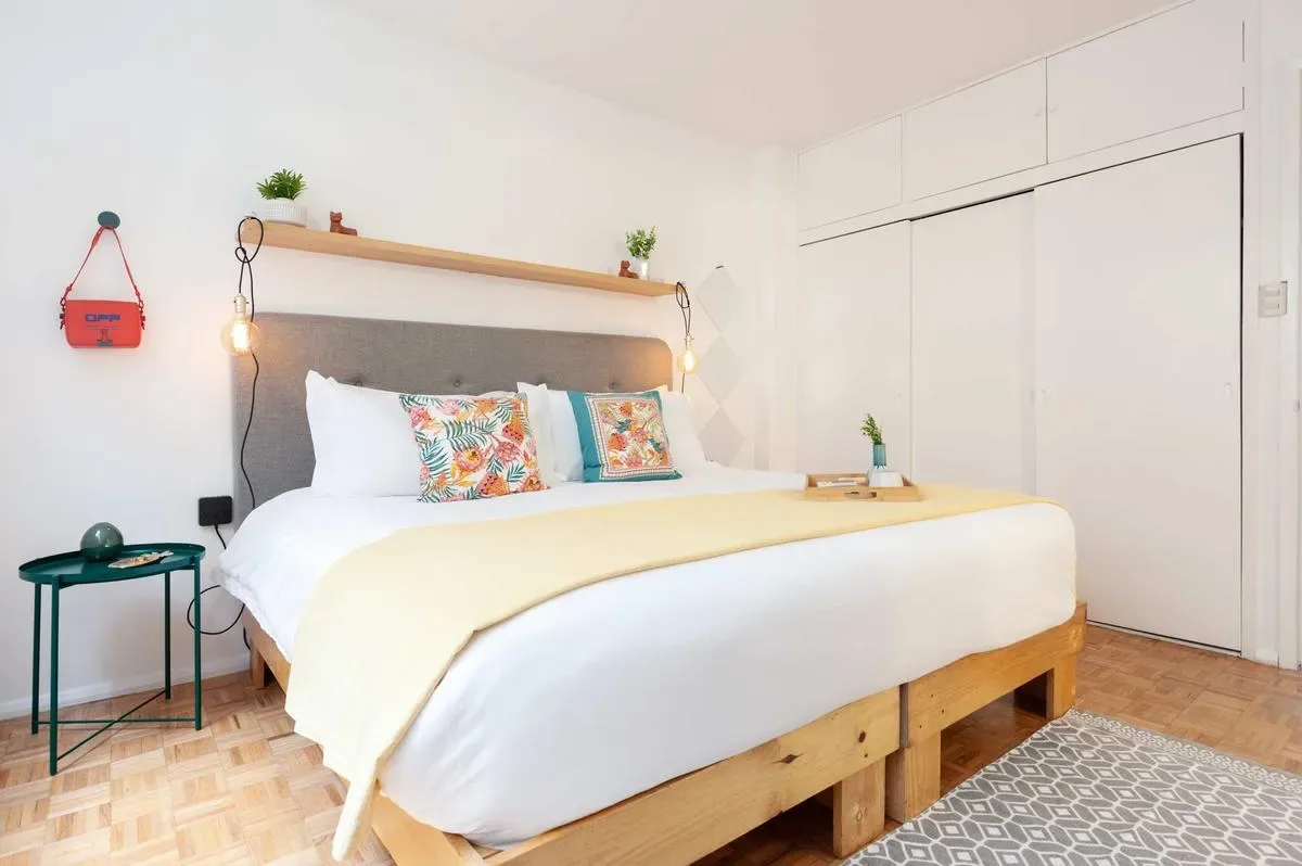 A bright bedroom features a wooden platform bed with colorful pillows, a bedside table, and a patterned rug.
