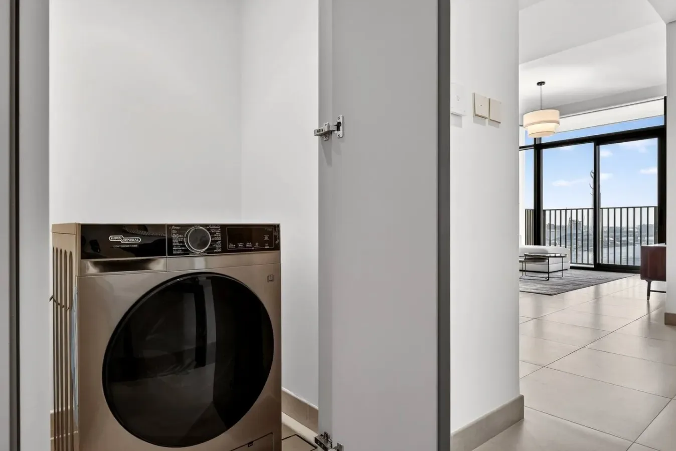 Zona de lavandería con una lavadora de carga frontal junto a una puerta abierta hacia una sala luminosa.