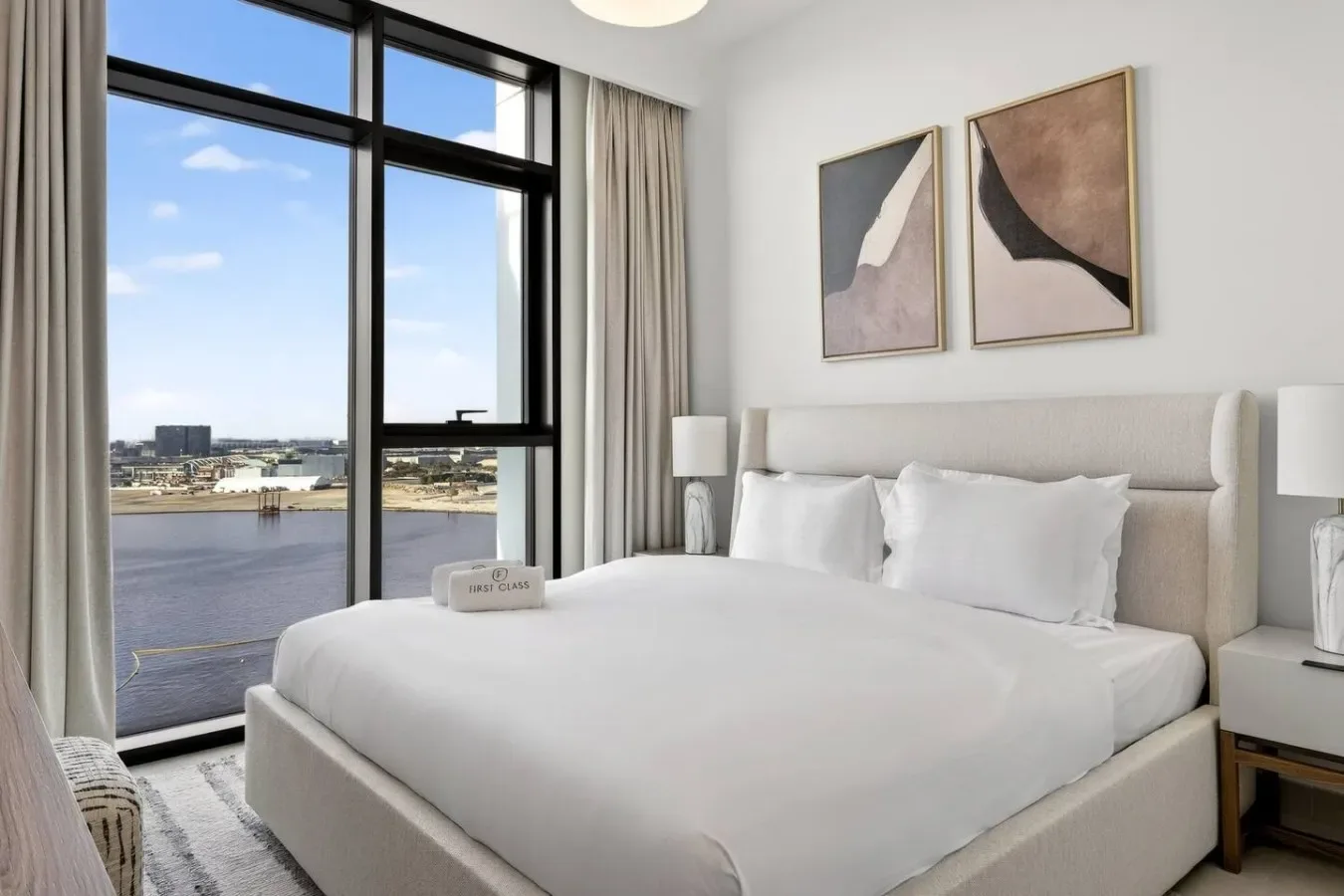 Un dormitorio luminoso con una cama tendida junto a grandes ventanas con vistas al agua.