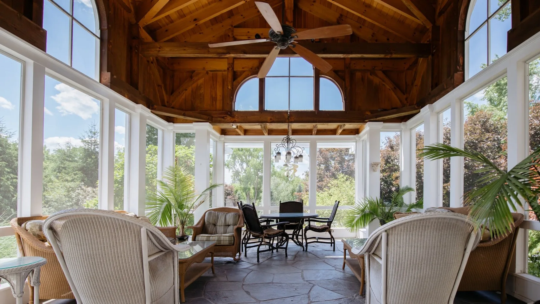A bright sunroom with seating and a dining table beneath a high wooden ceiling.