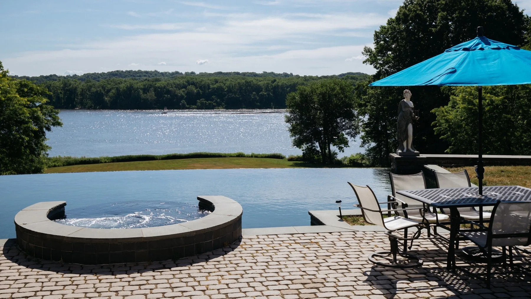 A lakeside patio features a circular hot tub, outdoor seating, and a blue umbrella.