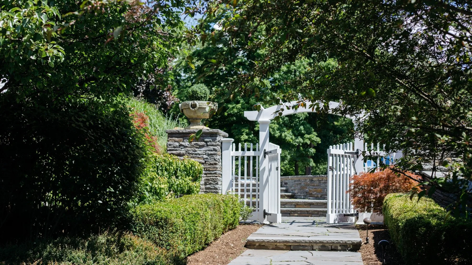 A garden walkway leads through an open white gate beneath leafy trees.