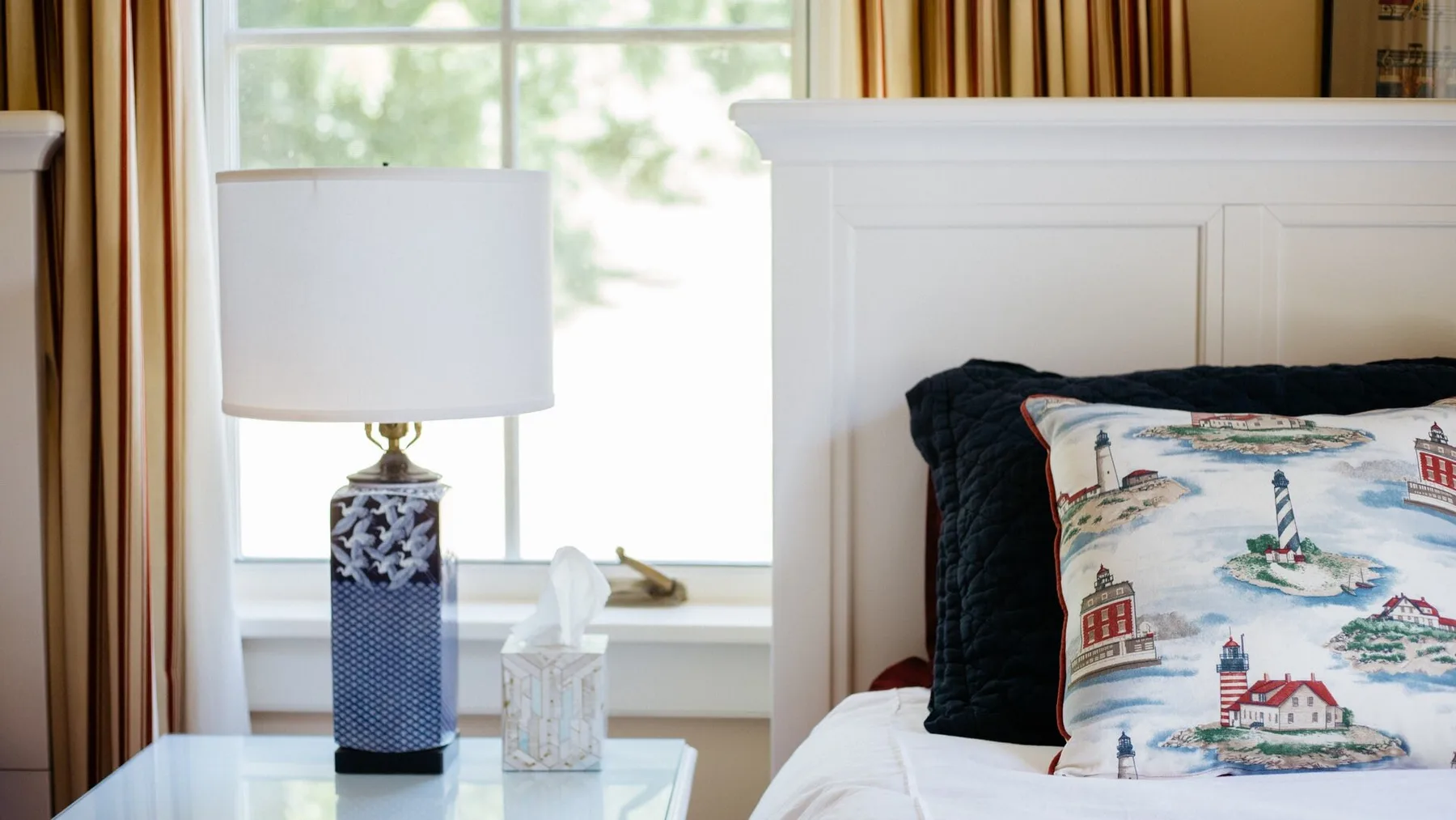 A bedroom with a white headboard, patterned pillows, and a bedside lamp.