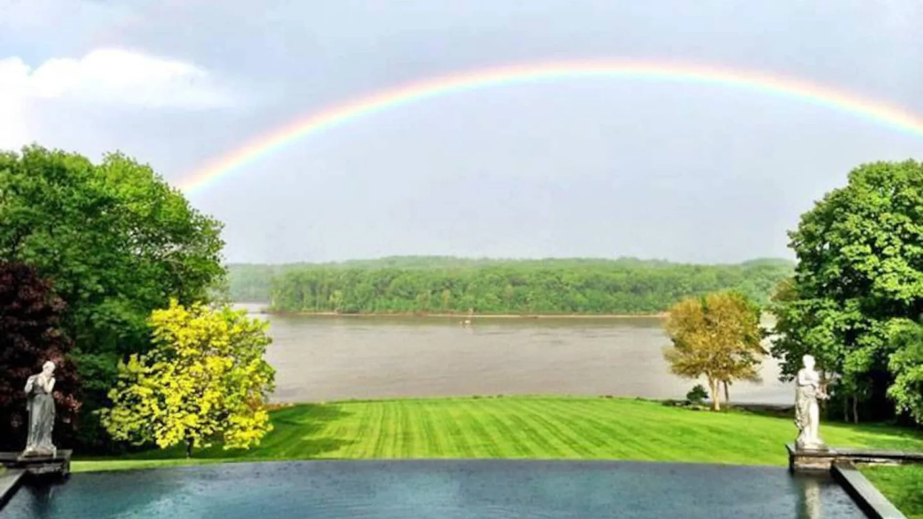 Un jardín cuidado bordea una piscina con vistas a un río bajo un arcoíris.