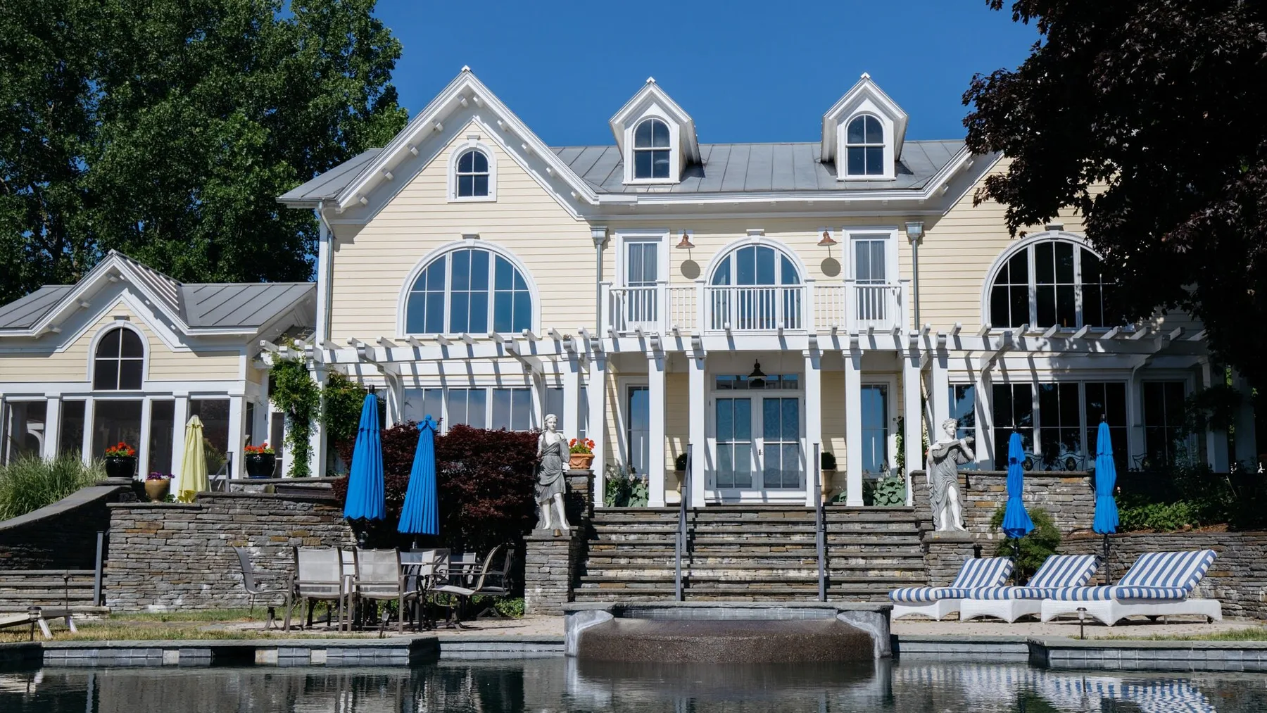 A large white house stands beside a swimming pool, with outdoor seating.