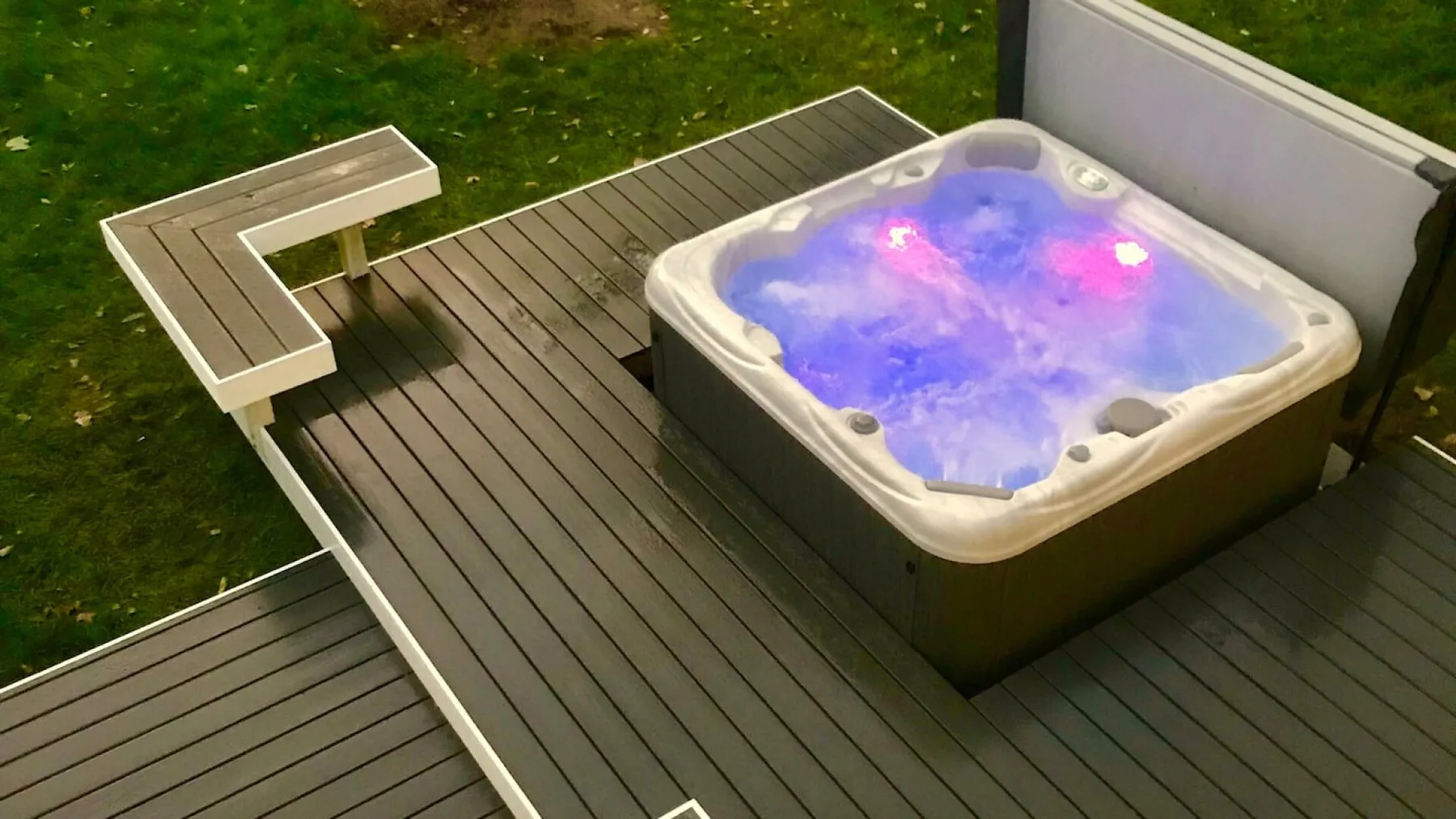 An outdoor hot tub glows beside a wooden deck and grassy area.
