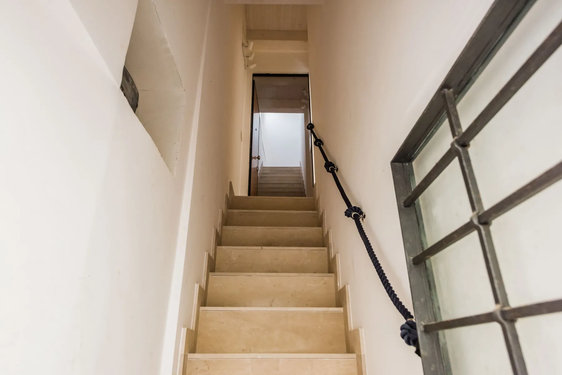 A narrow indoor staircase rises between white walls, with a dark handrail.