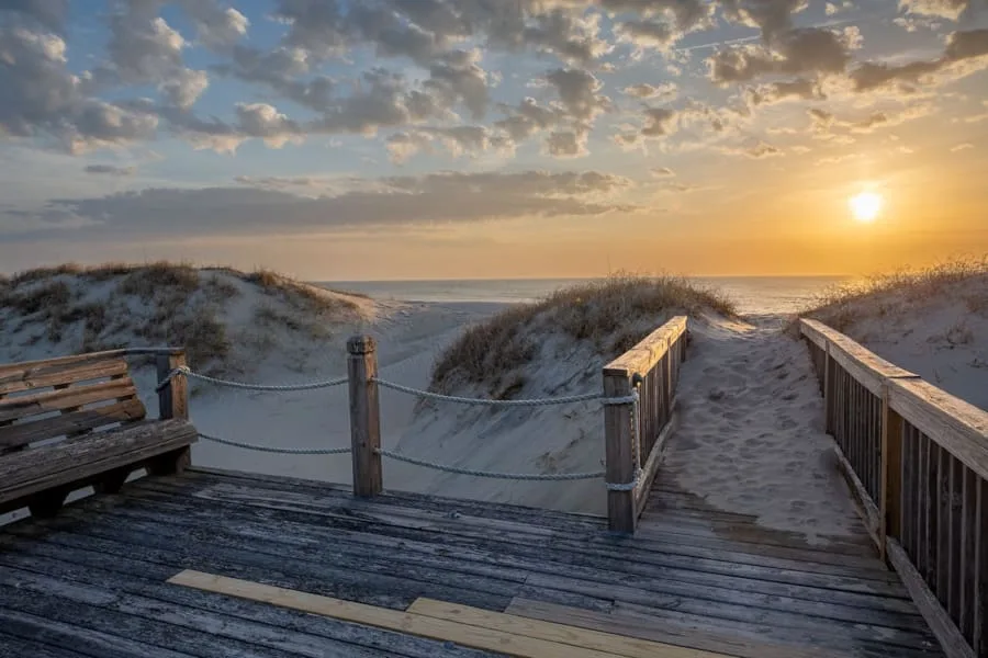 Una pasarela de madera cruza dunas de arena hacia el océano al atardecer.